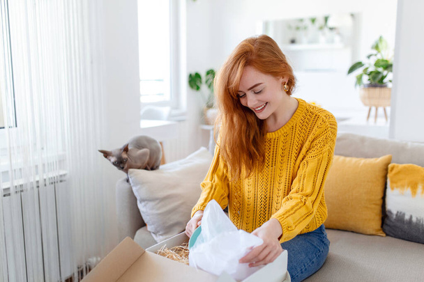 Χαμογελαστή γυναίκα κάθεται στον καναπέ στο σπίτι ανοικτό πακέτο μετά ψώνια σε απευθείας σύνδεση αγοράζοντας αγαθά στο διαδίκτυο, ευτυχισμένη νεαρή γυναίκα πελάτης ξεπακετάρει ταχυδρομικό δέμα αποστολής ικανοποιημένοι με την παραγγελία ή την παράδοση - Φωτογραφία, εικόνα