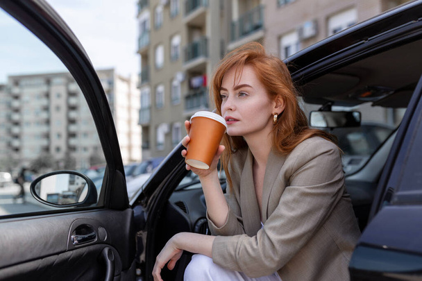 Ευτυχισμένη νεαρή γυναίκα με καφέ που έχει φρένο στο αμάξι της. Πλευρική άποψη της γυναίκας με καφέ για να πάει στο χέρι. Νεαρή γυναίκα πίνει καφέ στο αμάξι της. - Φωτογραφία, εικόνα
