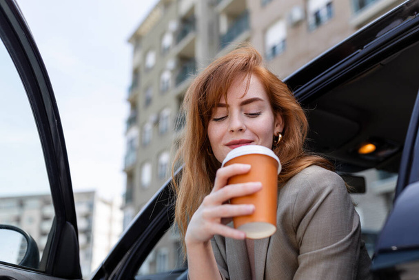 Jovencita feliz con café teniendo un freno en su coche. Vista lateral de mujer con café para ir en la mano. Mujer joven bebiendo café en su coche - Foto, imagen
