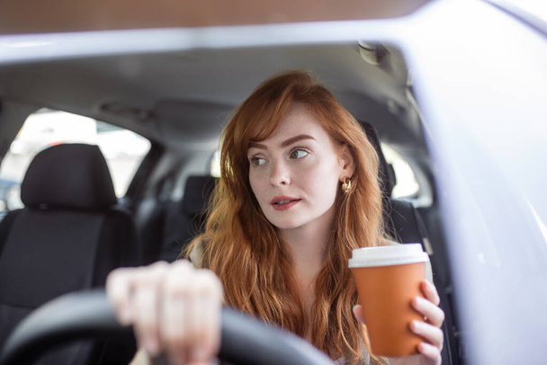 Mujer joven feliz con café para ir conduciendo su coche. Mujer bebiendo un café mientras conduce un coche. Mujer joven bebiendo café mientras conduce su coche. Atractivo pelo rojo conduce un coche - Foto, Imagen