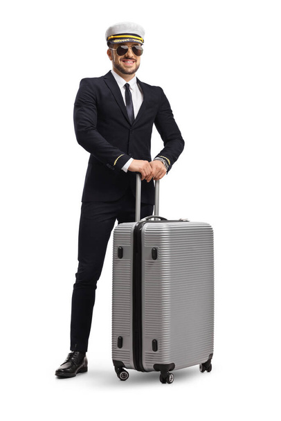 Young pilot in a uniform standing and holding a suitcase with wheels isolated on white background - Foto, afbeelding