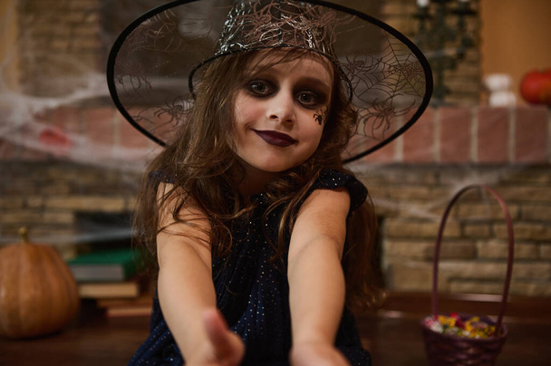 Όμορφο καυκάσιο κοριτσάκι με μακιγιάζ προσώπου - μια μαύρη αράχνη ζωγραφισμένη στο πρόσωπό της, που μοιάζει με μάγισσα ή μάγισσα, με καπέλο μάγου, έτοιμη για αποκριάτικο πάρτι. Γοτθικό στυλ. Φθινοπωρινό φεστιβάλ - Φωτογραφία, εικόνα