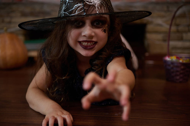 Little gothic girl, looking like an enchantress or witch in sorcerers hat reaches out her hand, staring at the camera with a frightening look, lying on the floor against a cobweb covered bricks wall - Photo, Image