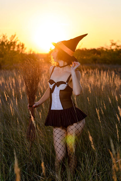 Sexy young woman in a witch's hat and a broom in a field, against the backdrop of a sunset. Concept for halloween. - Foto, Bild