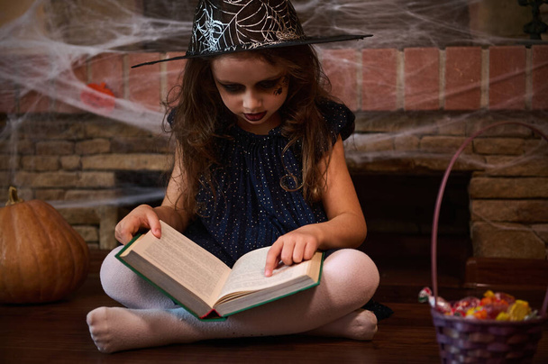 Gotická malá čarodějka v kouzelnickém klobouku, sedící na podlaze obklopená halloweenskými pochoutkami u pavučinami pokrytého krbu, seslala kouzla, zatímco četla knihu kouzel. Halloween party - Fotografie, Obrázek