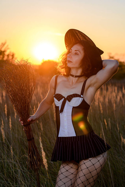 Sexy young woman in a witch's hat and a broom in a field, against the backdrop of a sunset. Concept for halloween. - Photo, Image