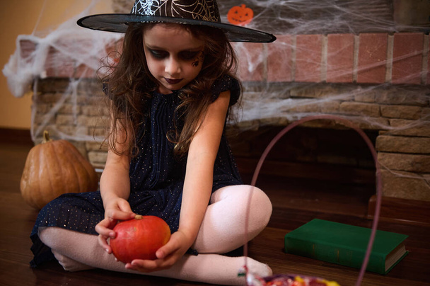 Adorable niña que parece una hechicera jugando con una calabaza naranja brillante, sentada en el suelo cerca de un libro de hechizos, contra una chimenea cubierta de telaraña. concepto de fiesta de Halloween - Foto, imagen