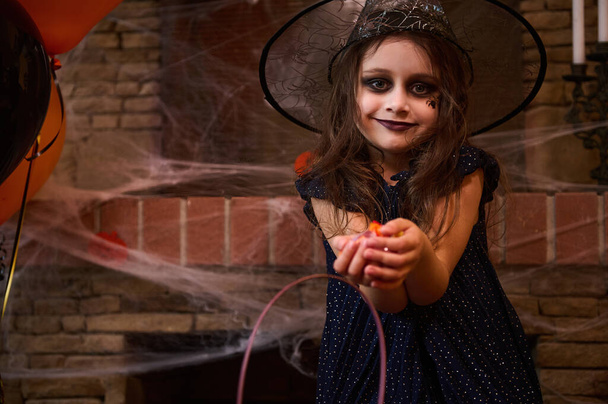 Biała słodka dziewczynka z zadymionym makijażem oczu, ubrana jak czarodziejka w kapeluszu czarodzieja, uśmiecha się patrząc w kamerę, trzymając garść cukierków, stojąc przed kominkiem pokrytym pajęczą pajęczyną - Zdjęcie, obraz