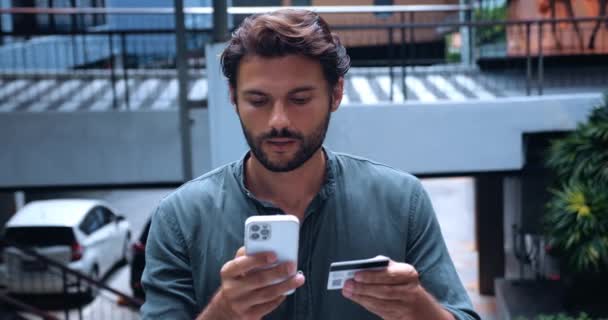 Atractivo joven caucásico hombre haciendo el pago en línea, utilizando el teléfono inteligente mientras se enfría en un acogedor café en un centro comercial - Imágenes, Vídeo