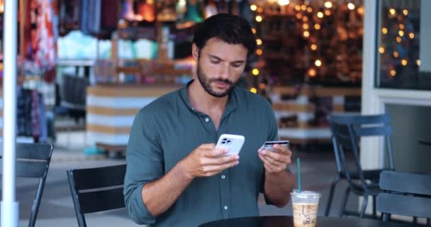 Atractivo hombre caucásico la transferencia de los datos de la tarjeta de crédito en el teléfono inteligente, mientras que hacer pagos sin efectivo en un café - Imágenes, Vídeo