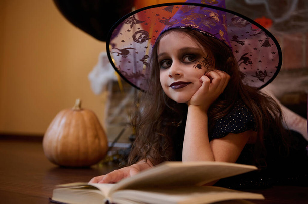 Criança adorável com uma aranha preta pintada na bochecha, parecendo uma feiticeira de chapéu de feiticeiro, sorri para a câmera enquanto estava deitada no chão cercada por um livro de feitiços, abóbora, decoração de Halloween - Foto, Imagem