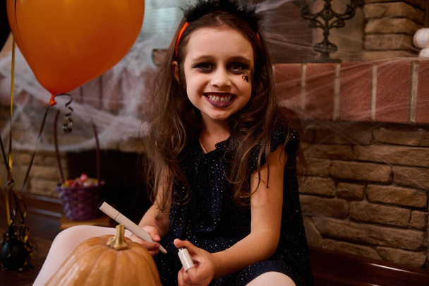Złośliwe dziecko, mała dziewczynka przebrana za wiedźmę z pomalowanym na twarzy pająkiem, uśmiechnięta do kamery, rysująca przerażającą twarz na dyni, Jack-O-Lantern. Koncepcja wesołego Halloween party - Zdjęcie, obraz