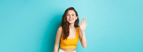 Summer holidays and emotions concept. Beautiful friendly woman with perfect body, waving hand and saying hi, greeting you, standing over blue background. - Photo, Image