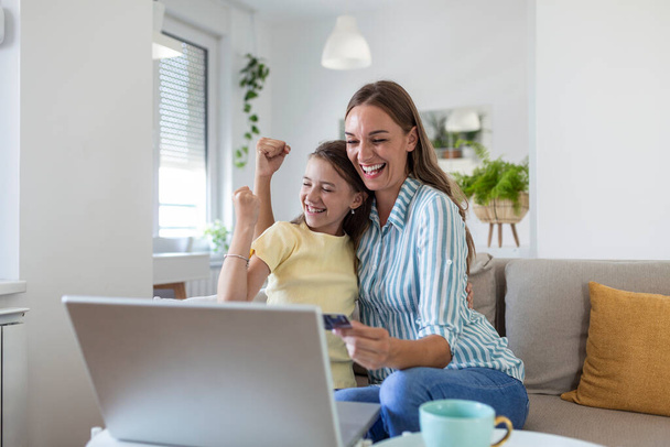 Low angle of happy small daughter with arms raised rejoicing while smiling mother in casual clothes focusing on screen and using laptop for online shopping in light modern apartment - Foto, imagen