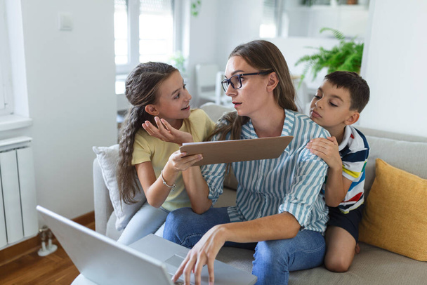 Frau arbeitet während der Quarantäne von zu Hause aus mit ihrem kleinen Sohn und ihrer Tochter auf der Couch und schreit und fordert Aufmerksamkeit - Foto, Bild
