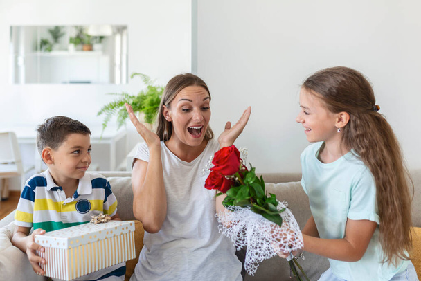Petite fille joyeuse avec bouquet de fleurs de roses et frère cadet avec boîte cadeau souriant et félicitant maman heureuse le jour de la fête des mères à la maison. Bonne fête des mères! - Photo, image