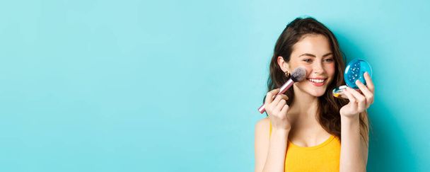Beauty. Modern girl with candid smile applying make up with brush, looking in pocket mirror, standing over blue background. - Foto, imagen