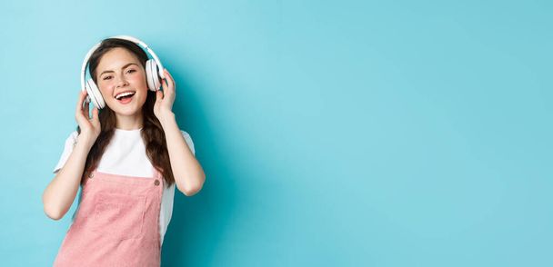Стильная современная девушка слушает музыку в беспроводных наушниках, трогает наушники и улыбается, поет вместе, стоя на синем фоне. - Фото, изображение
