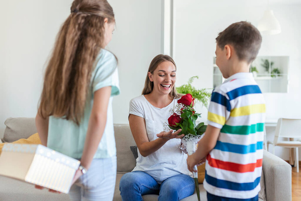 Счастливые дети дарят цветы матери. С Днем Матери! Дети мальчика и девочки поздравляют улыбающуюся мать, дарят ей букет цветов из роз и подарочную коробку во время праздника - Фото, изображение