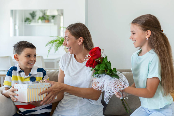 Веселая маленькая девочка с букетом цветов из роз и младший брат с подарочной коробкой улыбается и поздравляет счастливую маму с днем матери дома. С Днем матери!! - Фото, изображение