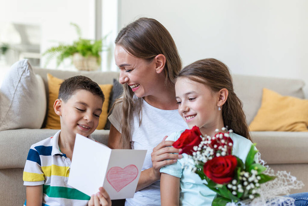 Νεαρή μητέρα με μπουκέτο τριαντάφυλλα γελάει, αγκαλιάζει το γιο της, και; heerful κορίτσι με μια κάρτα και τριαντάφυλλα συγχαίρει τη μαμά κατά τη διάρκεια εορτασμού των διακοπών στο σπίτι. Ημέρα μητέρων - Φωτογραφία, εικόνα