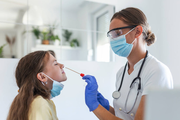 Enfermera realizando una prueba de hisopo nasal a un niño pequeño. Chica pasando por pruebas de PCR debido a la pandemia de COVID-19. Médico femenino usando hisopo de algodón mientras prueba de PCR niña pequeña - Foto, imagen