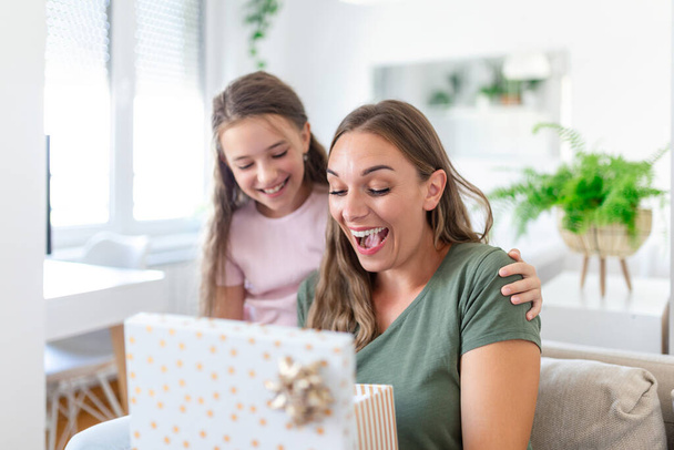 Junge Frau und Mädchen zu Hause feiern Muttertag sitzend auf Sofa Tochter umarmt Mutter küsst Wange Mama lacht freudig halten Geschenkbox - Foto, Bild