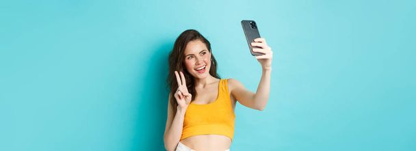 テクノロジーとライフスタイルの概念。若い女性が夏の服装で自撮りし、スマートフォンのカメラでvサインを見せ、青い背景の上に立つ. - 写真・画像