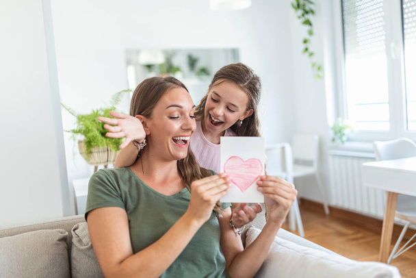 Junge Frau und Mädchen zu Hause feiern Muttertag sitzend auf Sofa Tochter umarmt Mutter küsst Wange Mama lacht freudig halten Geschenkbox - Foto, Bild