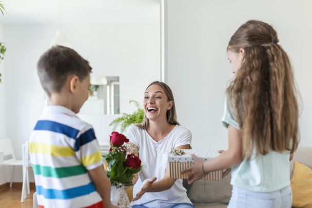 Cheerful little girl with gift box and youngest brother with bouquet of roses flowers smiling and congratulating happy mom on mother day at home. Happy Mothers Day! - Photo, Image