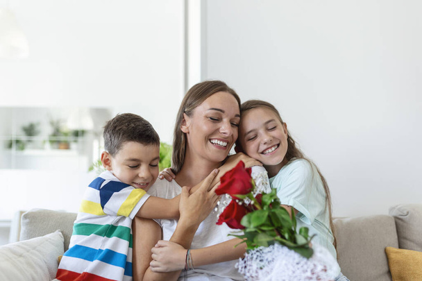 Młoda matka z bukietem róż śmieje się, przytula syna, a? wesoła dziewczyna z kartką gratuluje mamie podczas świąt w kuchni w domu - Zdjęcie, obraz