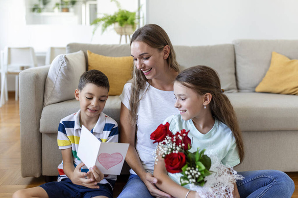 Молодая мама с букетом роз смеется, обнимает сына, и "прелестная девочка с открыткой и розами поздравляет маму во время праздника дома. День матери - Фото, изображение