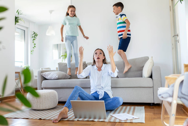 Уставшая молодая мама сидит на полу и работает с ноутбуком и документами, пока маленькие дети и прыгать на диване и веселиться и шуметь - Фото, изображение