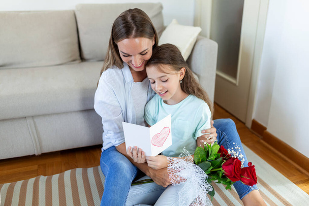 Šťastný Den matek! Dcera gratuluje mámě a dává jí pohlednici a květiny. Máma s holkou se usmívají a objímají. Rodinná dovolená a sounáležitost. - Fotografie, Obrázek