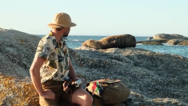 A мужчина путешественник в сафари шляпе разбился лагерь и покоится на камне рядом с океаном, и наблюдает за спящим меховым тюленем. мужчина в отпуске в Южной Африке фотографирует тюленя на пляже - Кадры, видео
