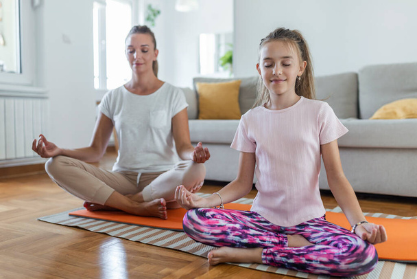 Yoga con niños. Joven madre de familia e hija pequeña meditando con los ojos cerrados mientras se sienta en la pose de loto en la flor en la sala de estar en casa - Foto, Imagen
