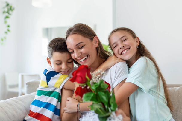 Νεαρή μητέρα με ένα μπουκέτο τριαντάφυλλα γελάει, αγκαλιάζει το γιο της, και; heerful κορίτσι με μια κάρτα συγχαίρει τη μαμά κατά τη διάρκεια εορτασμού διακοπών στην κουζίνα στο σπίτι - Φωτογραφία, εικόνα