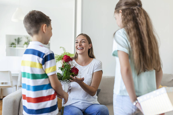 Счастливые дети дарят цветы матери. С Днем Матери! Дети мальчика и девочки поздравляют улыбающуюся мать, дарят ей букет цветов из роз и подарочную коробку во время праздника - Фото, изображение