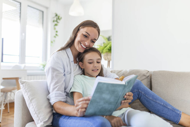 Loving young mother reading book to adorable little daughter, sitting on cozy sofa in living room, mum teaching preschool girl child, family spending weekend at home together, children education - Photo, Image