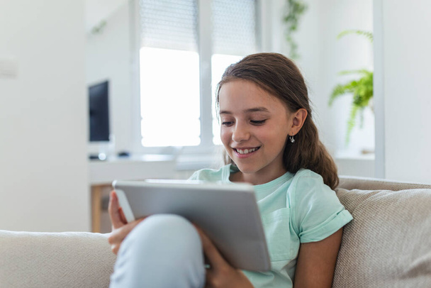 Evdeki oturma odasında dijital tablet pedini kullanarak kanepede oturan tatlı küçük mutlu kız gülüşü. Aile etkinliği kavramı. - Fotoğraf, Görsel