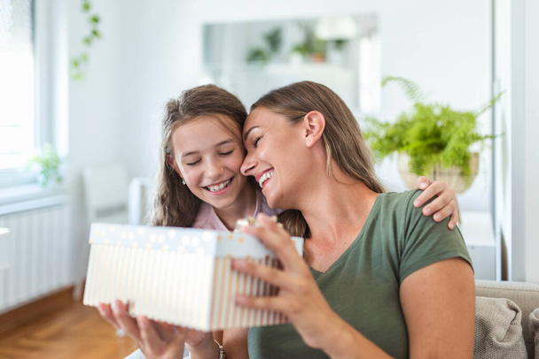 Young woman and girl at home celebrating mother's day sitting on sofa daughter hugging mother kissing cheek mom laughing joyful holding gift box - 写真・画像