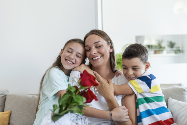 バラの花束を持つ若い母親は笑い、彼女の息子を抱き、そして?家でのキッチンでの休日のお祝い中にカードを持つ相続人の女の子はお母さんを祝います - 写真・画像