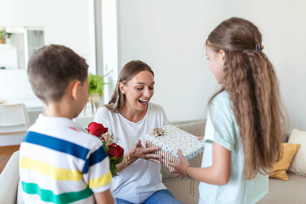 Szczęśliwe dzieci dają matce kwiaty. Szczęśliwego Dnia Matki! Dzieci chłopiec i dziewczyna gratulacje uśmiechnięta matka, dać jej bukiet kwiatów róż i pudełko prezentów podczas uroczystości wakacyjnych - Zdjęcie, obraz
