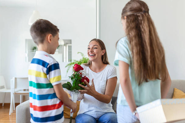 Happy children giving gift an flowers to mother. Happy Mothers Day! Children boy and girl congratulate smiling mother, give her flowers bouquet of roses and a gift box during holiday celebration - Photo, Image