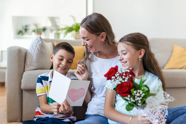Jeune mère avec un bouquet de roses rit, étreignant son fils, et ? heerful fille avec une carte félicite maman pendant la célébration des vacances dans la cuisine à la maison - Photo, image