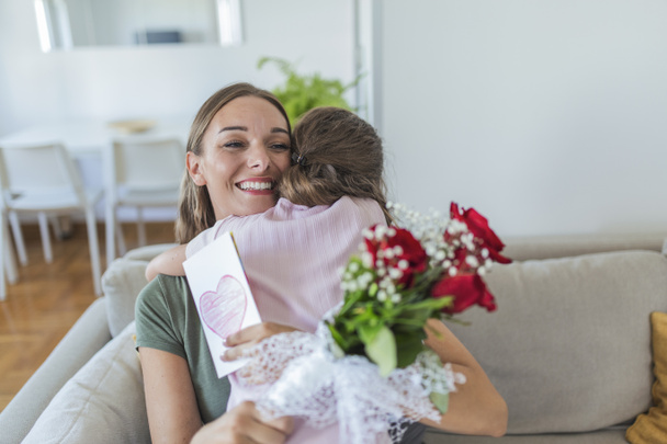 Αγαπώ τη μαμά μου! Ελκυστική νεαρή γυναίκα με μικρό χαριτωμένο κορίτσι περνούν χρόνο μαζί στο σπίτι, ευχαριστώντας για χειροποίητη κάρτα με σύμβολο αγάπης και λουλούδια. Χαρούμενη οικογενειακή ιδέα. Ημέρα της Μητέρας. - Φωτογραφία, εικόνα