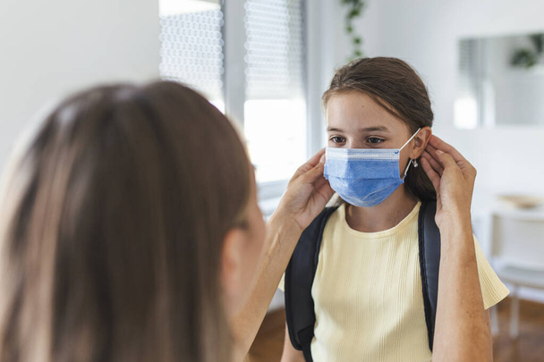Genç anne okula hazırlanmak için kızının tıbbi maske takmasına yardım ediyor. Covid-19 ya da koronavirüs salgınından kaçınıyorum. Okula geri dön. - Fotoğraf, Görsel