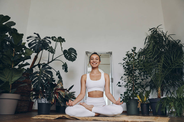 Happy young woman meditating while sitting on the floor with houseplants all around her - Foto, imagen