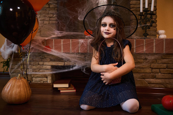 Hezká holčička, čarodějka v kouzelnickém klobouku a kouřovém make-upu, objímající košík halloweenských lahůdek, sedící na podlaze obklopená knihami kouzel a Jack-O-Lanternem, u krbu s pavučinou - Fotografie, Obrázek