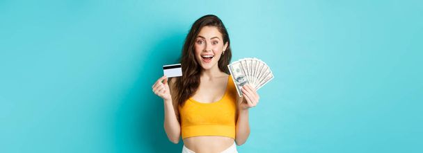 Yazlık kıyafetli çekici bir kadın, para ve plastik kredi kartı gösteriyor, şaşırmış bir şekilde gülümsüyor, mavi arka planda duruyor.. - Fotoğraf, Görsel